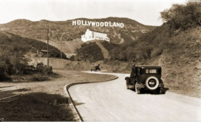 An old photo of a car driving down a road with the hollywood sign in the background.
