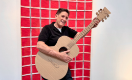 A man holding a cardboard guitar in front of a red wall.